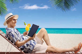 Old man sitting on beach chair by ocean reading a book and sipping on a cocktail
